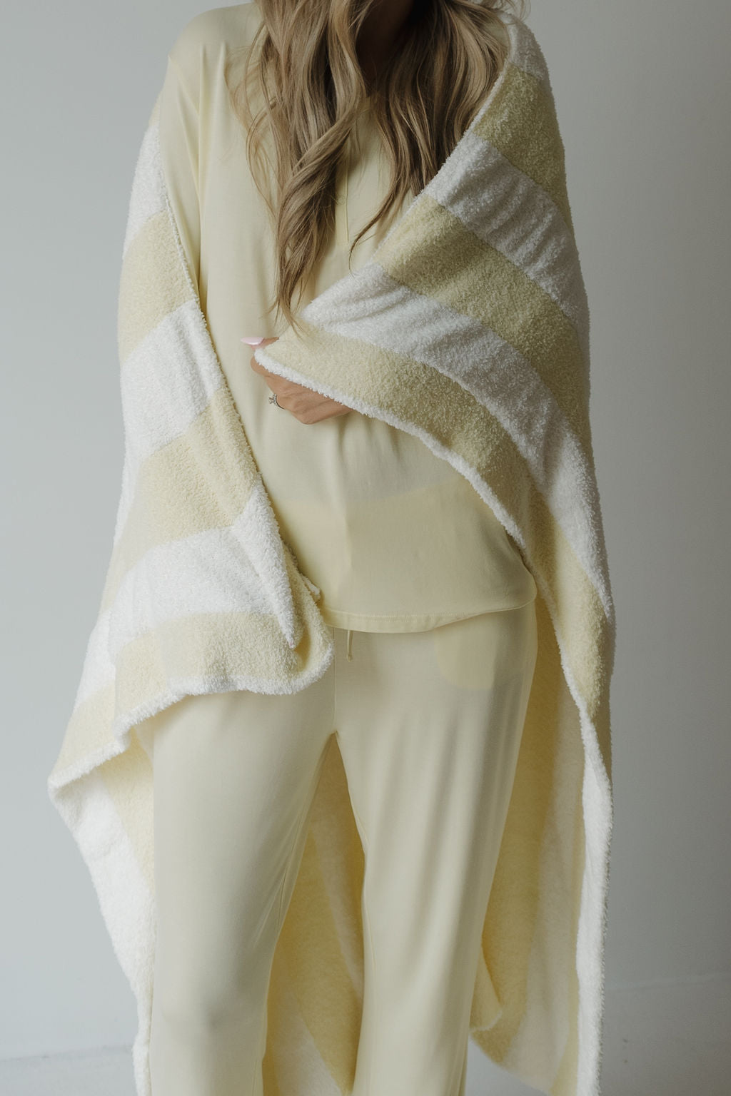 A person with long, wavy hair is wearing the Women’s Long Sleeve Bamboo Pajama in Sunny by forever french baby—breastfeeding friendly and light yellow—wrapped in a soft, striped yellow and white blanket, standing against a plain light background.