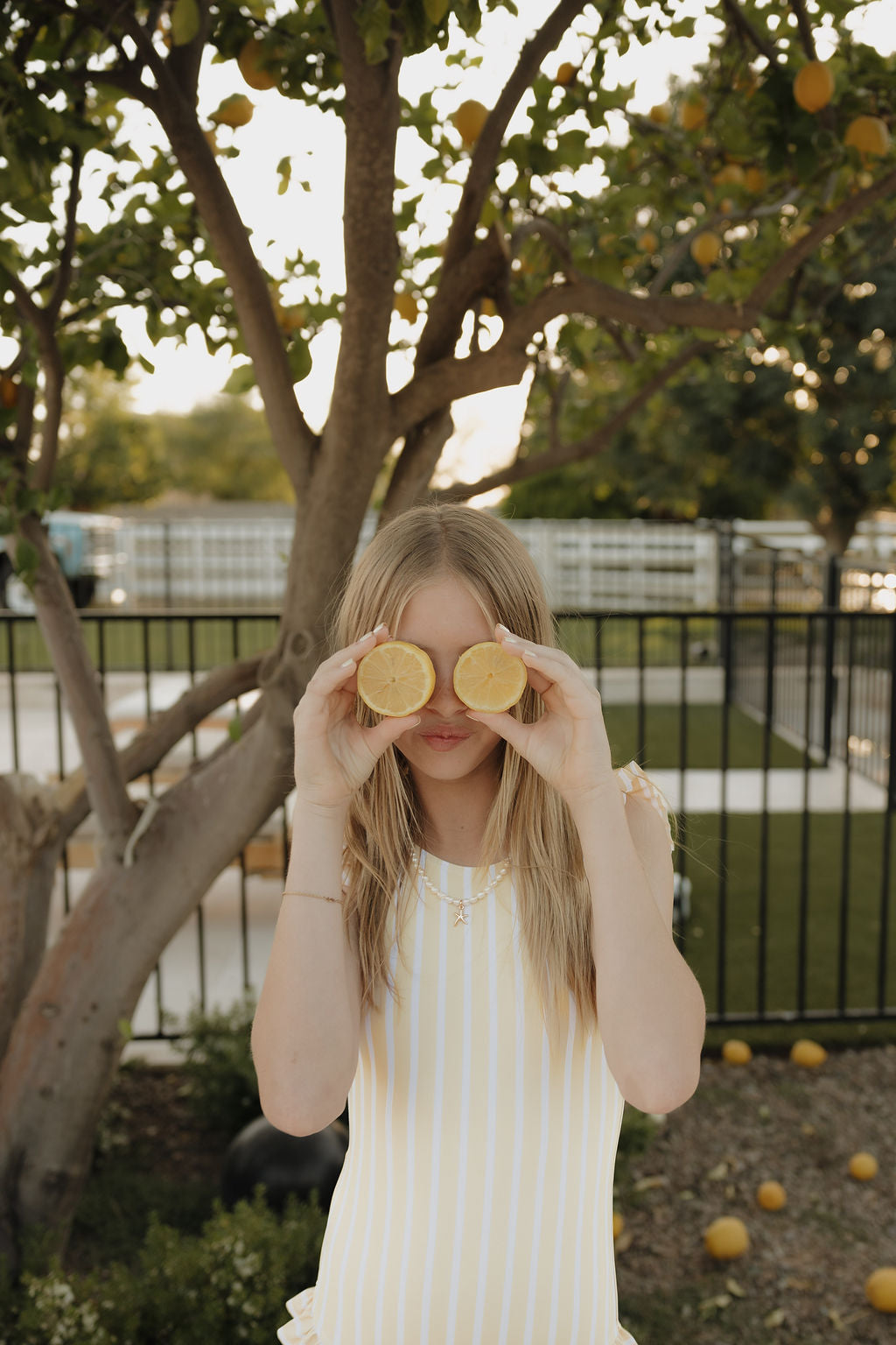 A girl wearing the lolo webb 2 Girl Sleeveless Swimsuit | Sun Rays playfully poses outdoors, holding lemon halves over her eyes in front of a leafy lemon tree with fruit.