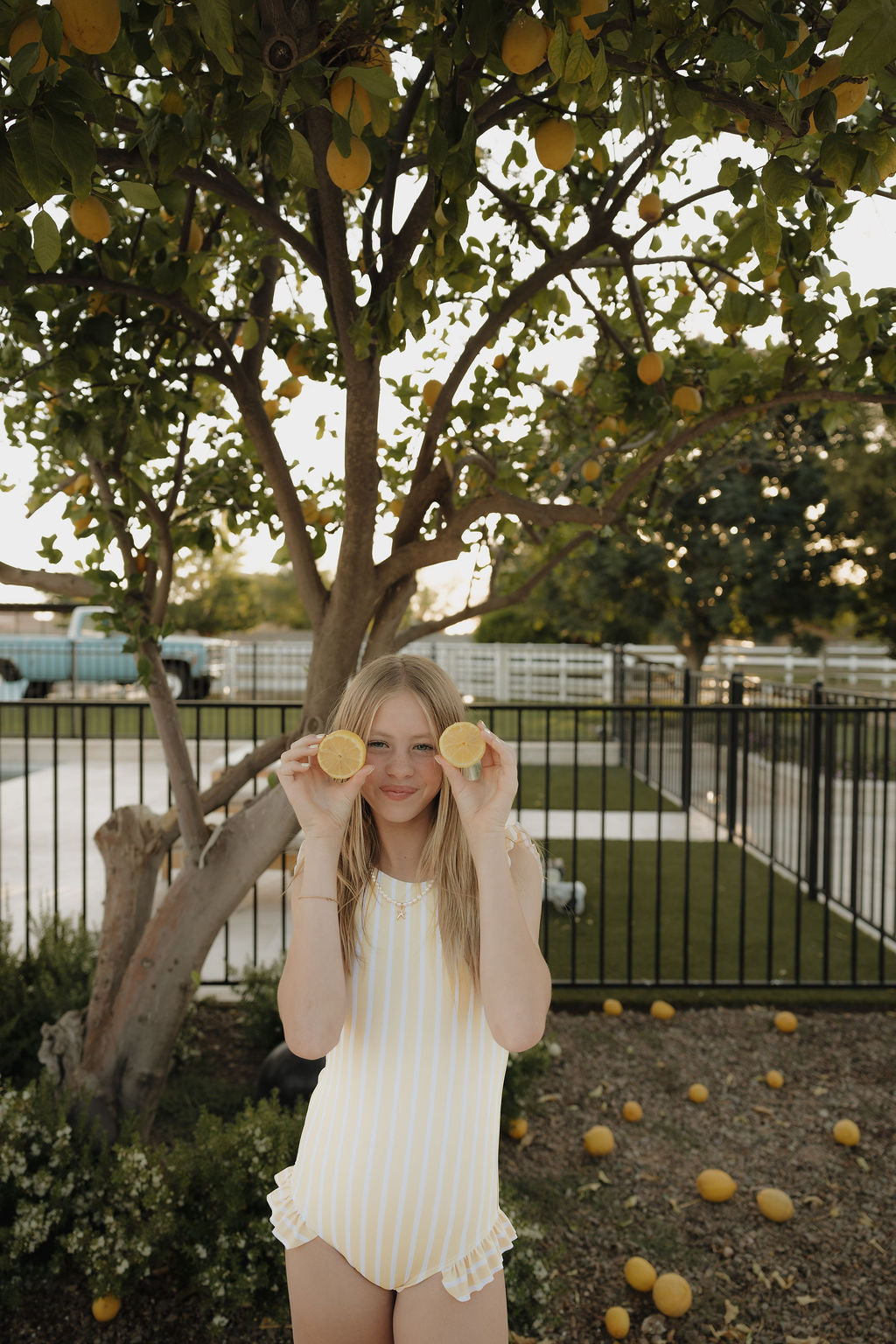 A girl wears the lolo webb 2 Girl Sleeveless Swimsuit | Sun Rays with yellow and white stripes, stands under a lemon tree and holds lemon halves over her eyes, smiling. Lemons are scattered on the ground with a fence in the background.