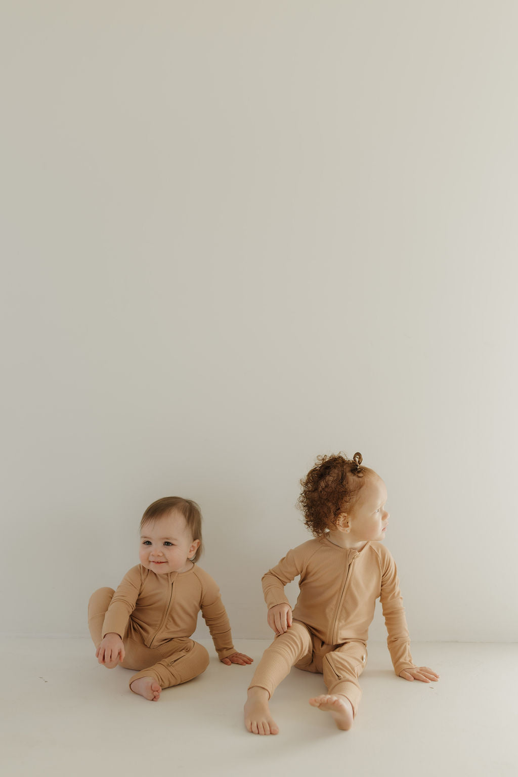 Two young children dressed in Forever French Baby's Bamboo Zip Pajamas in Tawny sit on a light floor against a plain wall. One child sits upright, looking ahead, while the other looks to the side. Their light expressions reflect the comfort of breathable babywear.