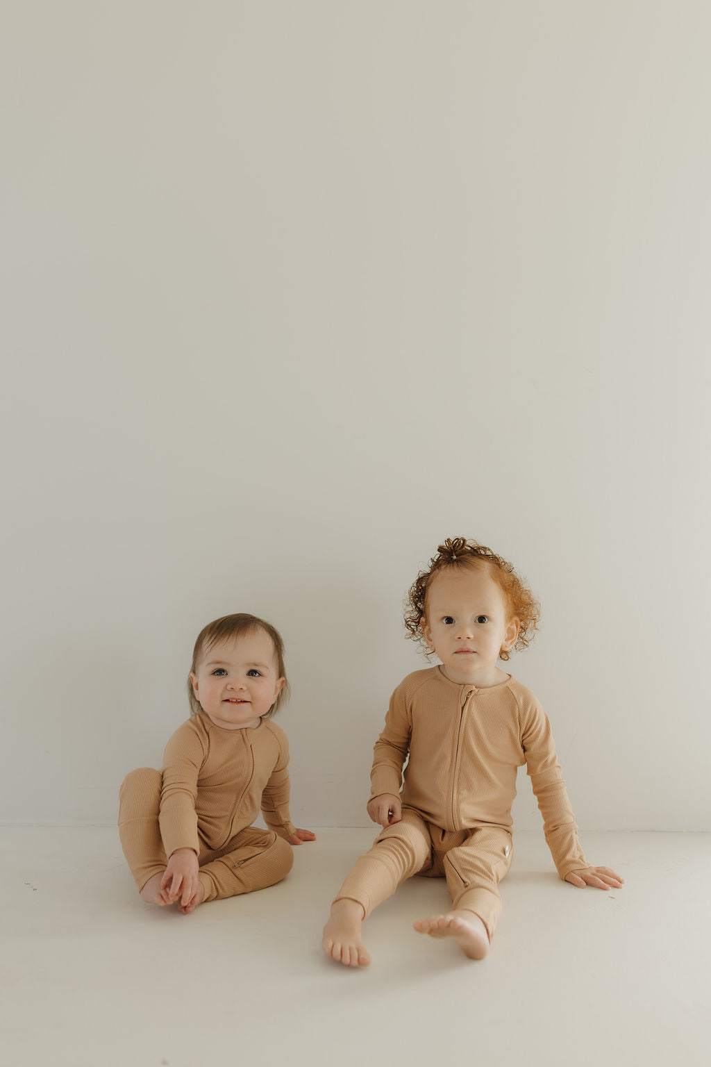 Two toddlers sit against a white background wearing forever french baby's Bamboo Zip Pajamas in Tawny. One has light brown hair and smiles, while the other has curly hair and a neutral expression, both enjoying the comfort of breathable babywear.