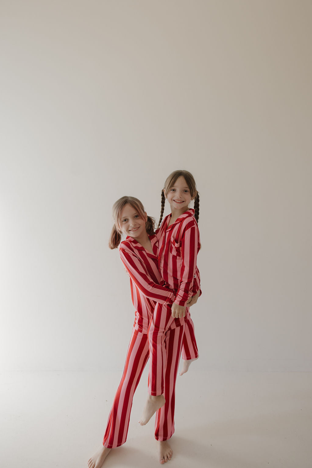 Two young girls wearing Minty Cherries' Toddler Bamboo Button Up Lounge Set in Iconic Stripe stand barefoot and smiling in a minimal room, enjoying the cozy, hypo-allergenic comfort of their matching lounge sets.