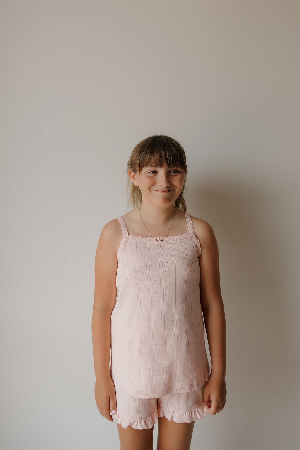 A young girl with light brown hair and bangs smiles while standing against a plain light-colored wall, wearing the forever french baby Girl’s Ruffle Short Set | Ballerina.