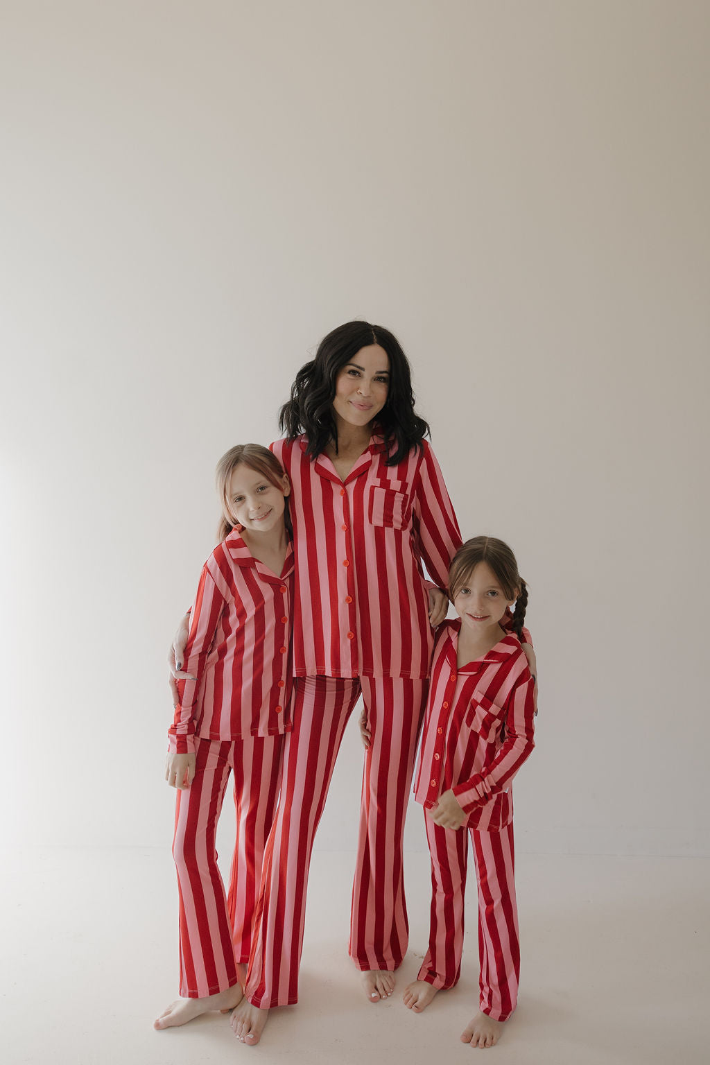 A woman and two girls stand barefoot, smiling in matching Minty Cherries Toddler Bamboo Button Up Lounge Set | Iconic Stripe—adorable, hypo-allergenic pajamas that make comfort and style look effortless against a light backdrop.