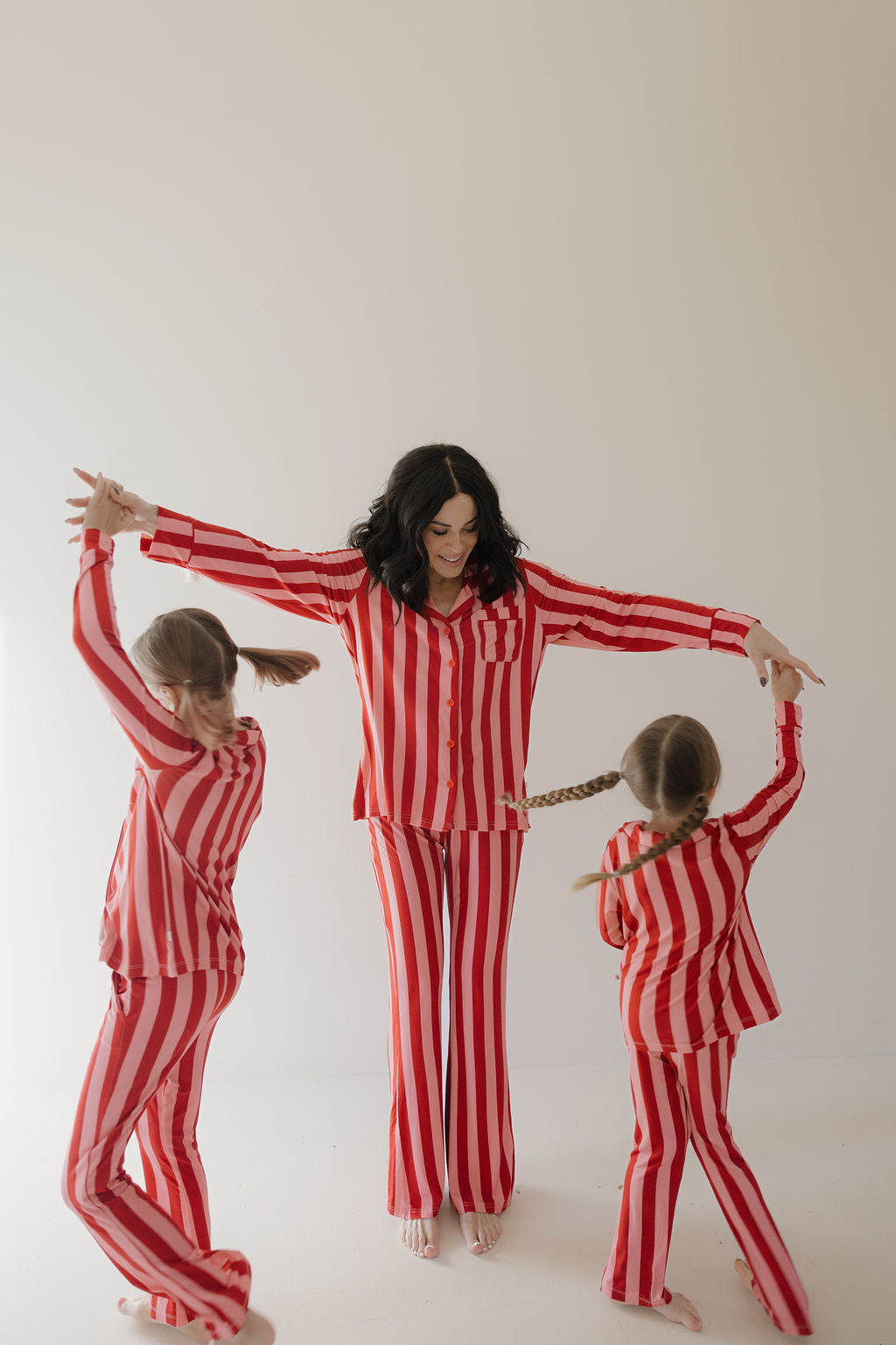 A woman and two young girls, all wearing Minty Cherries Women's Bamboo Button Up Lounge Set in Iconic Stripe, joyfully dance together in a bright, minimal room.