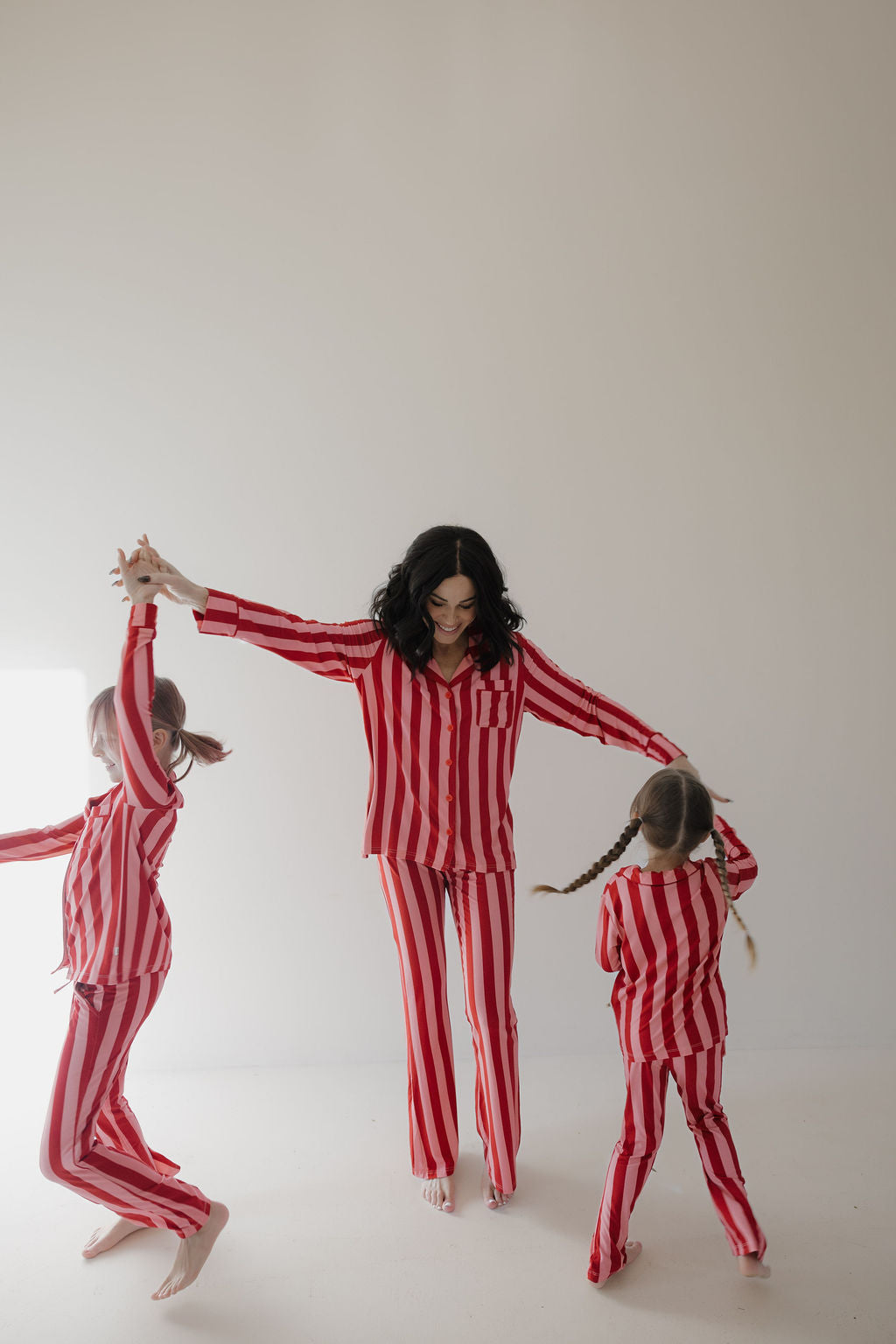 A woman and two children wear matching Minty Cherries Toddler Bamboo Button Up Lounge Sets in Iconic Stripe as they hold hands and dance in a bright, minimal room, enjoying the comfort of soft, hypo-allergenic bamboo fabric.