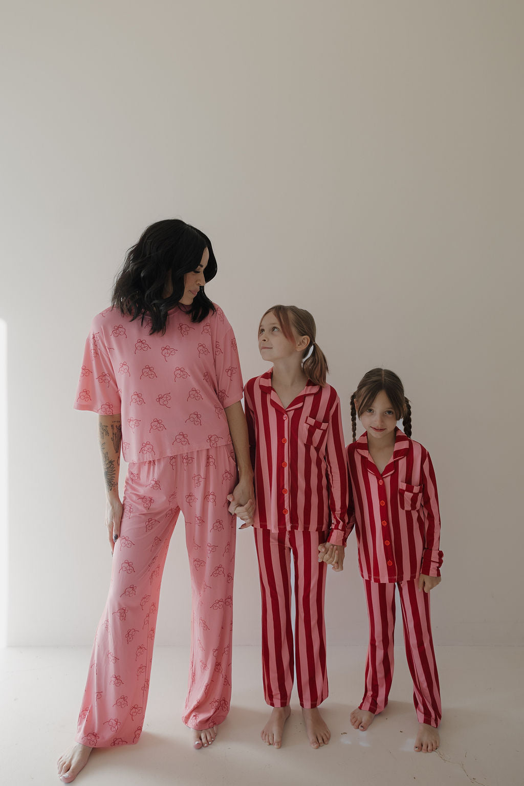 A woman in a Minty Cherries Women's Boxy Tee Lounge Set in Iconic Cherry stands barefoot, holding hands with two girls in matching red-and-pink striped loungewear, against a plain light background, all looking at each other.