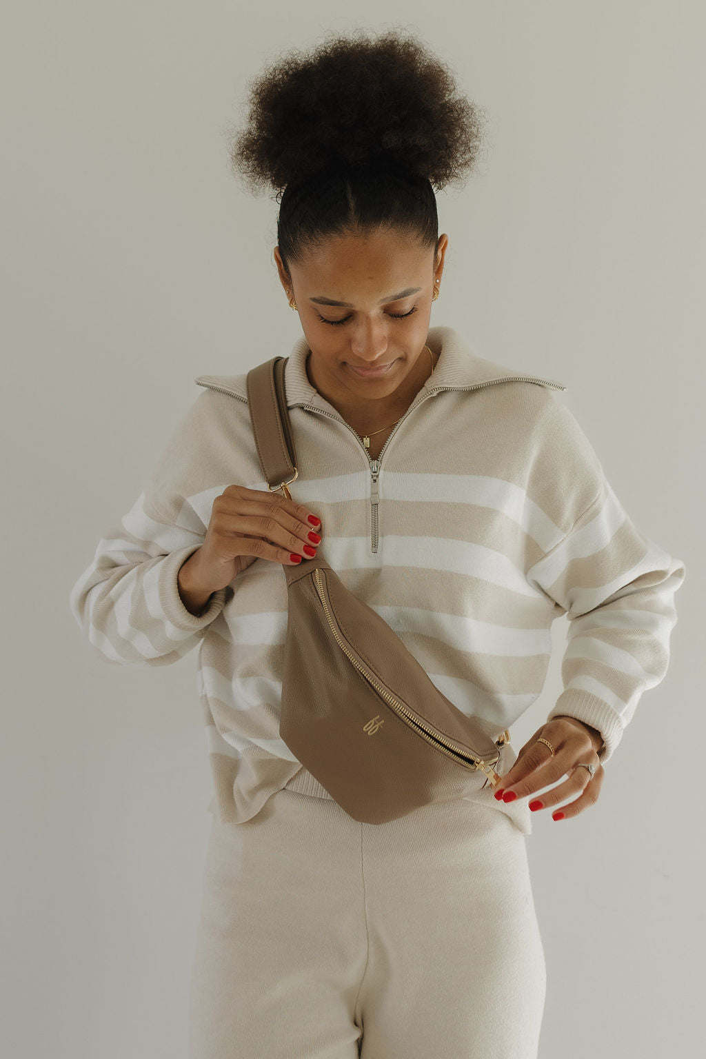 A person is adjusting a chic ff Belt Bag in Taupe by forever french baby, while wearing a beige and white striped sweater and cream pants. With curly hair and red nail polish, they stand gracefully against a neutral background.