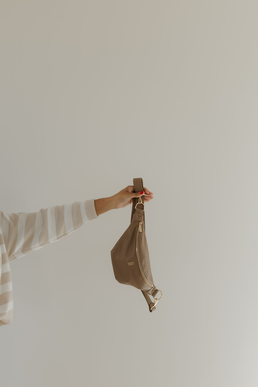 A person wearing a striped sweater holds up a taupe ff Belt Bag from Forever French Baby against a neutral background. It features a gold zipper and hardware, offering chic style in every detail.