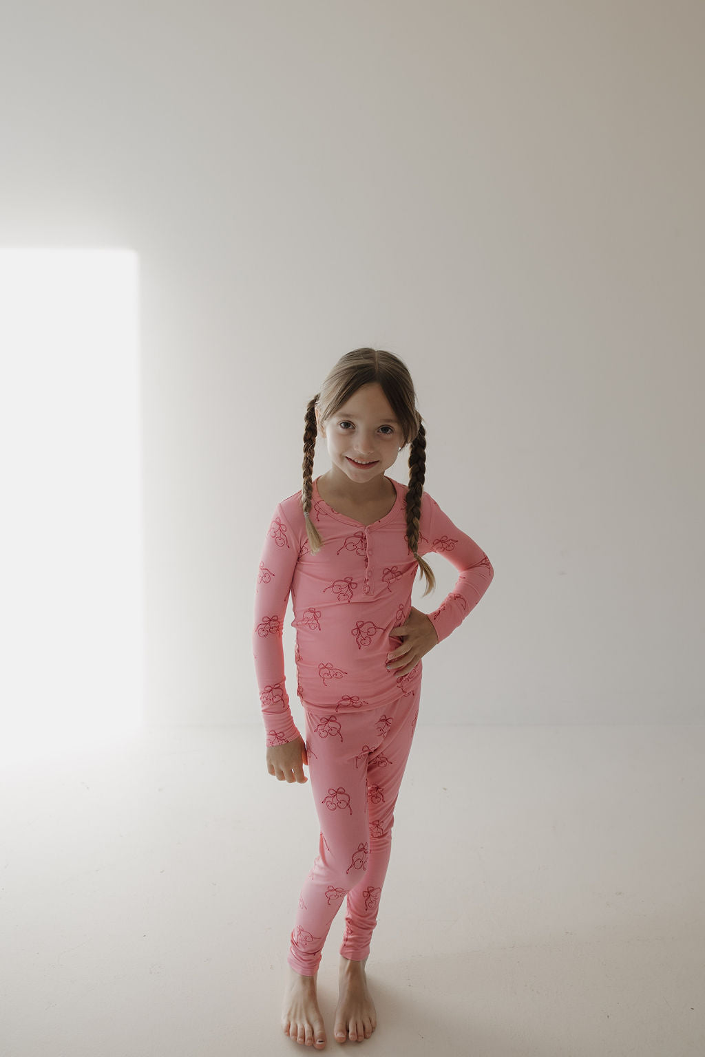 A young girl with braided hair stands barefoot in a minimalist room, smiling in Minty Cherries' Bamboo Two Piece Pajamas | Iconic Cherry. Hand on her hip, she enjoys the breathable comfort as light streams through the window.