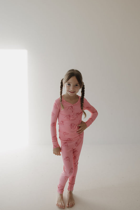 A young girl with braided hair stands barefoot in a minimalist room, smiling in Minty Cherries' Bamboo Two Piece Pajamas | Iconic Cherry. Hand on her hip, she enjoys the breathable comfort as light streams through the window.