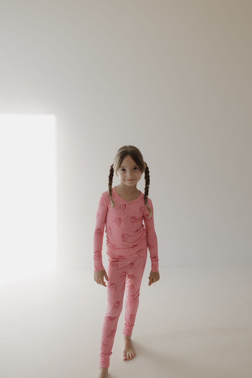 A young girl with brown braided hair stands barefoot on a white floor, smiling slightly at the camera. She wears Minty Cherries’ Bamboo Two Piece Pajamas in Iconic Cherry print. The background is a plain light wall with a bright window.