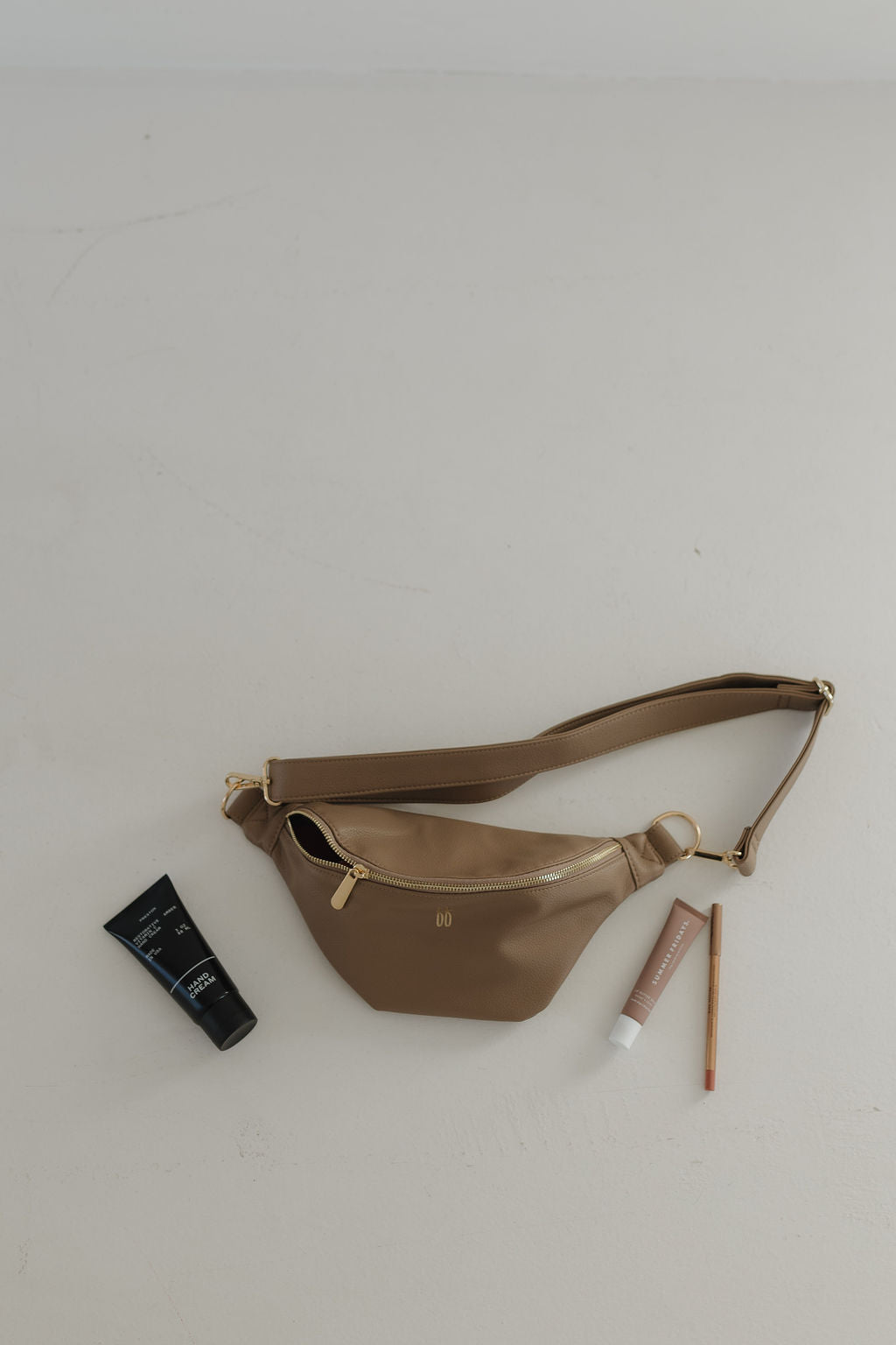 A taupe faux leather belt bag by Forever French Baby is showcased on a light surface with a gold zipper and strap, next to a black tube, beige tube, and pencil-like cosmetic item.