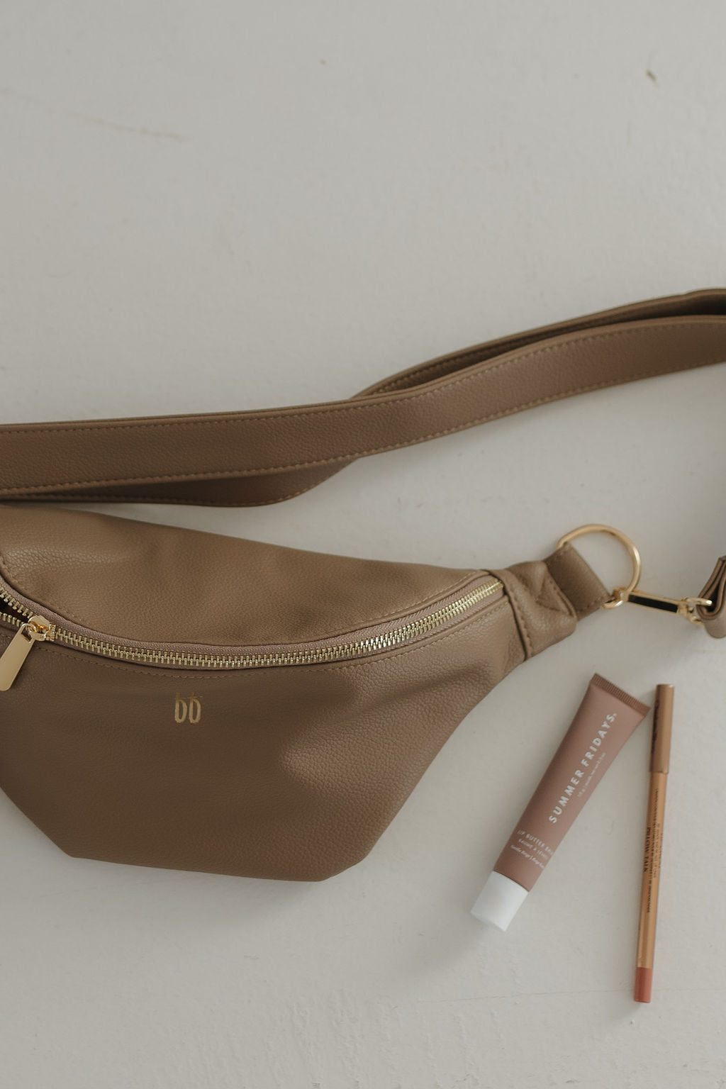 A taupe ff Belt Bag from forever french baby, featuring a simple strap and gold zipper, rests on a light surface with a tube of cream and a lip liner.