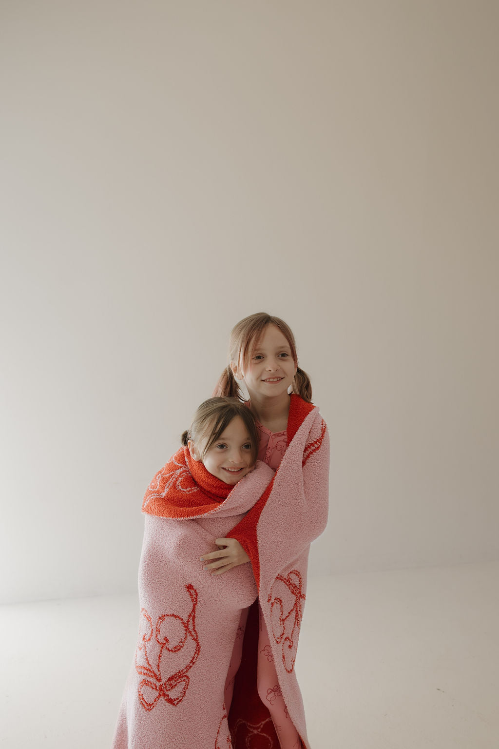 Two young girls with light brown hair smile and hug while wrapped in the Minty Cherries Plush Blanket | Iconic Cherry, featuring cartoon designs, in a bright room with a plain light-colored background.