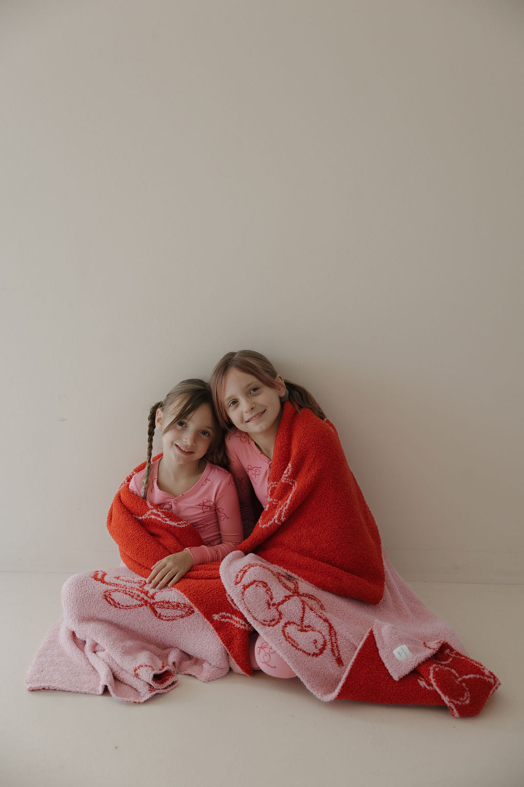 Two young girls with brown hair sit smiling on the floor in Minty Cherries' Bamboo Two Piece Pajamas | Iconic Cherry, wrapped in cozy, breathable red and pink sleepwear for a cheerful, comfortable look.