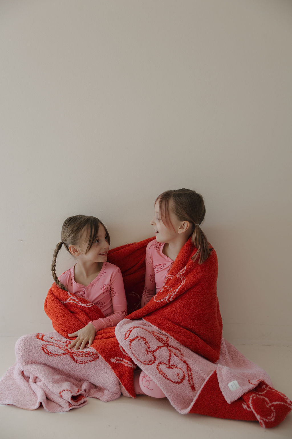 Two young girls in matching pink pajamas sit on the floor, smiling at each other while wrapped in the Minty Cherries Plush Blanket | Iconic Cherry. Both have braided brown hair. The background is a plain, light-colored wall.
