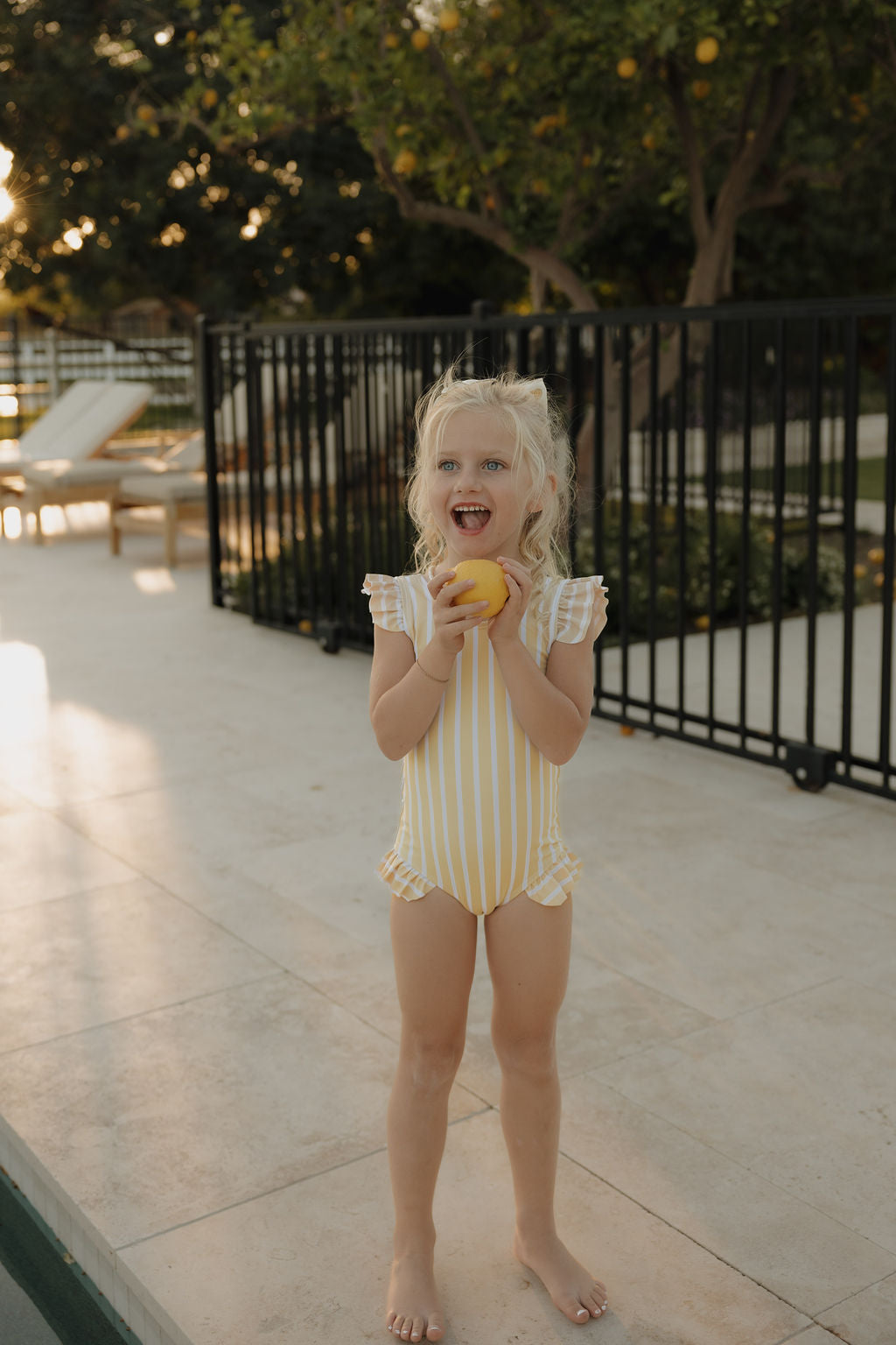A blonde girl smiles poolside in the lolo webb 2 Girl Sleeveless Swimsuit | Sun Rays, holding a lemon. Trees and a black metal fence are in the background—perfect summer style.