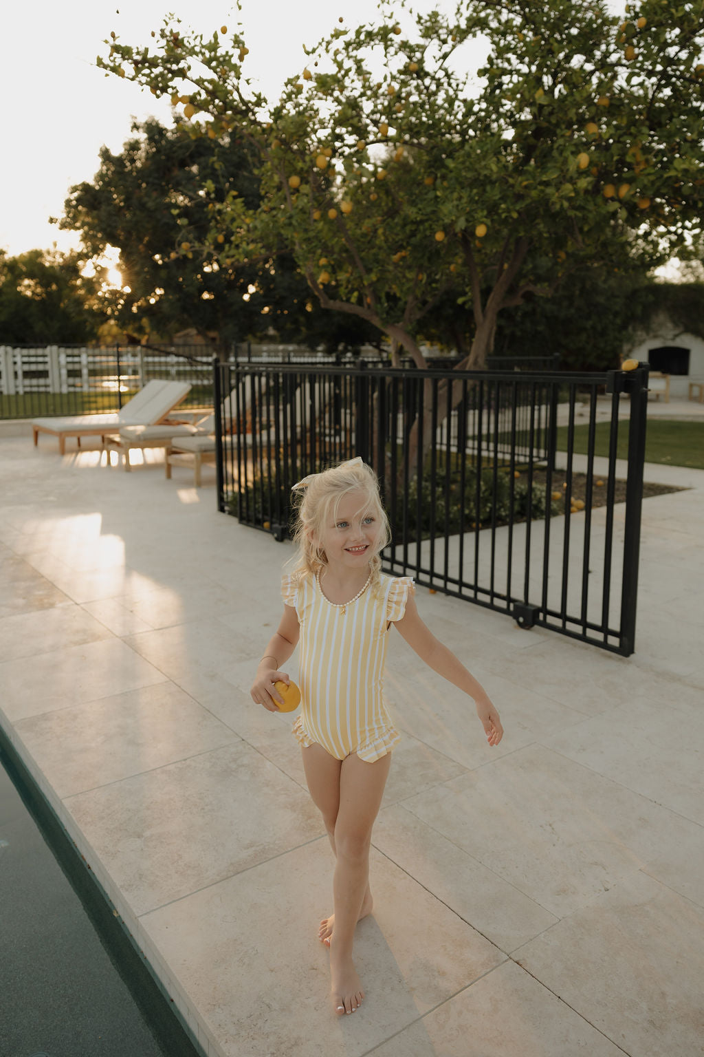 A young girl smiles by the pool, barefoot and holding a yellow ball, wearing the lolo webb 2 Girl Sleeveless Swimsuit | Sun Rays with yellow and white stripes and ruffles; trees and lounge chairs are in the background.