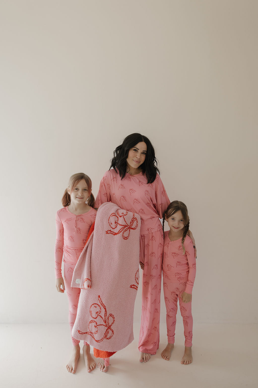 A woman and two girls wear matching Minty Cherries Women's Boxy Tee Lounge Sets in Iconic Cherry print, holding a pink cherry blanket—cozy, hypo-allergenic bamboo pajamas ideal for family nights.