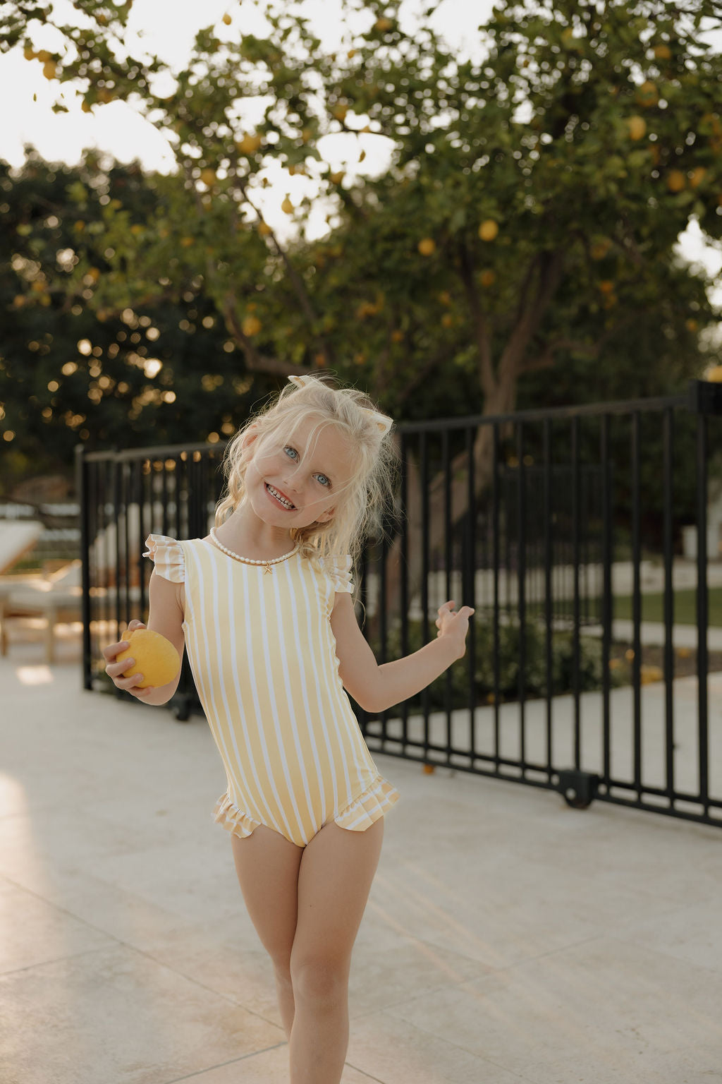 A young blonde girl smiles and holds a lemon outdoors in warm sunlight, wearing the lolo webb 2 Girl Sleeveless Swimsuit | Sun Rays, featuring yellow and white stripes with charming ruffle detail for forever French summer vibes.