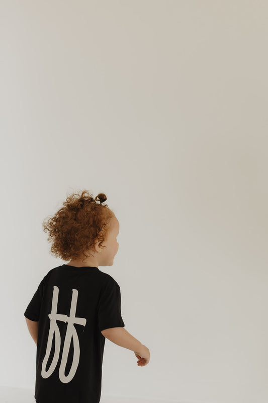 A toddler with curly hair wears the forever french baby Toddler Short Sleeve Bamboo Lounge Set in Black ff Signature, featuring white symbols on the back. The child faces left, walking against a plain, light background.