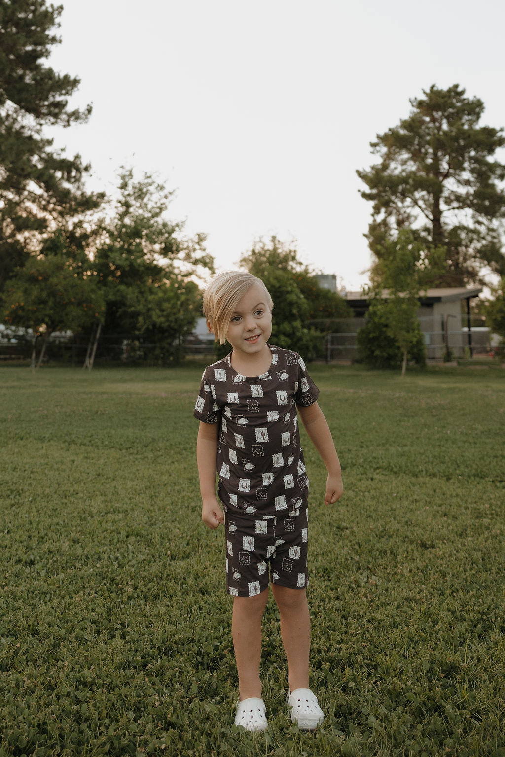 A young child with light hair stands on a grassy lawn, wearing the Bamboo Short Set by forever french baby. White shoes complete the look. In the background are trees and a building under a clear sky.