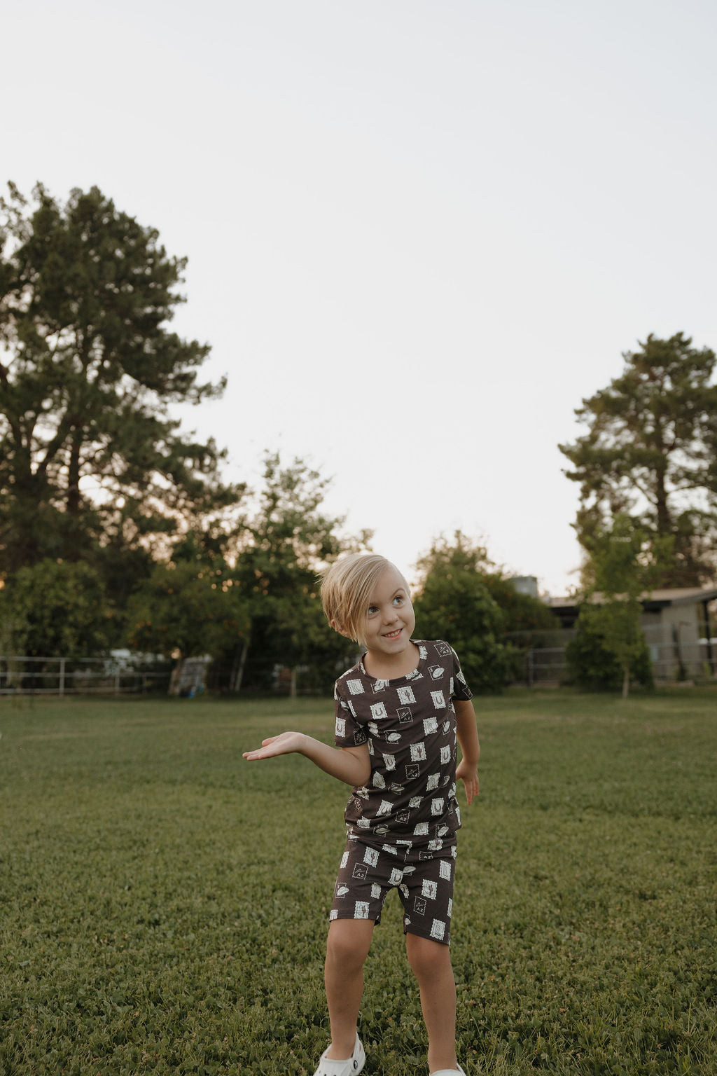 A young blonde child stands on grass smiling, playfully reaching out while wearing the forever french baby Bamboo Short Set | With Love from AZ—featuring dark bamboo fabric with light patterns. Trees and a building appear under a clear sky in the background.
