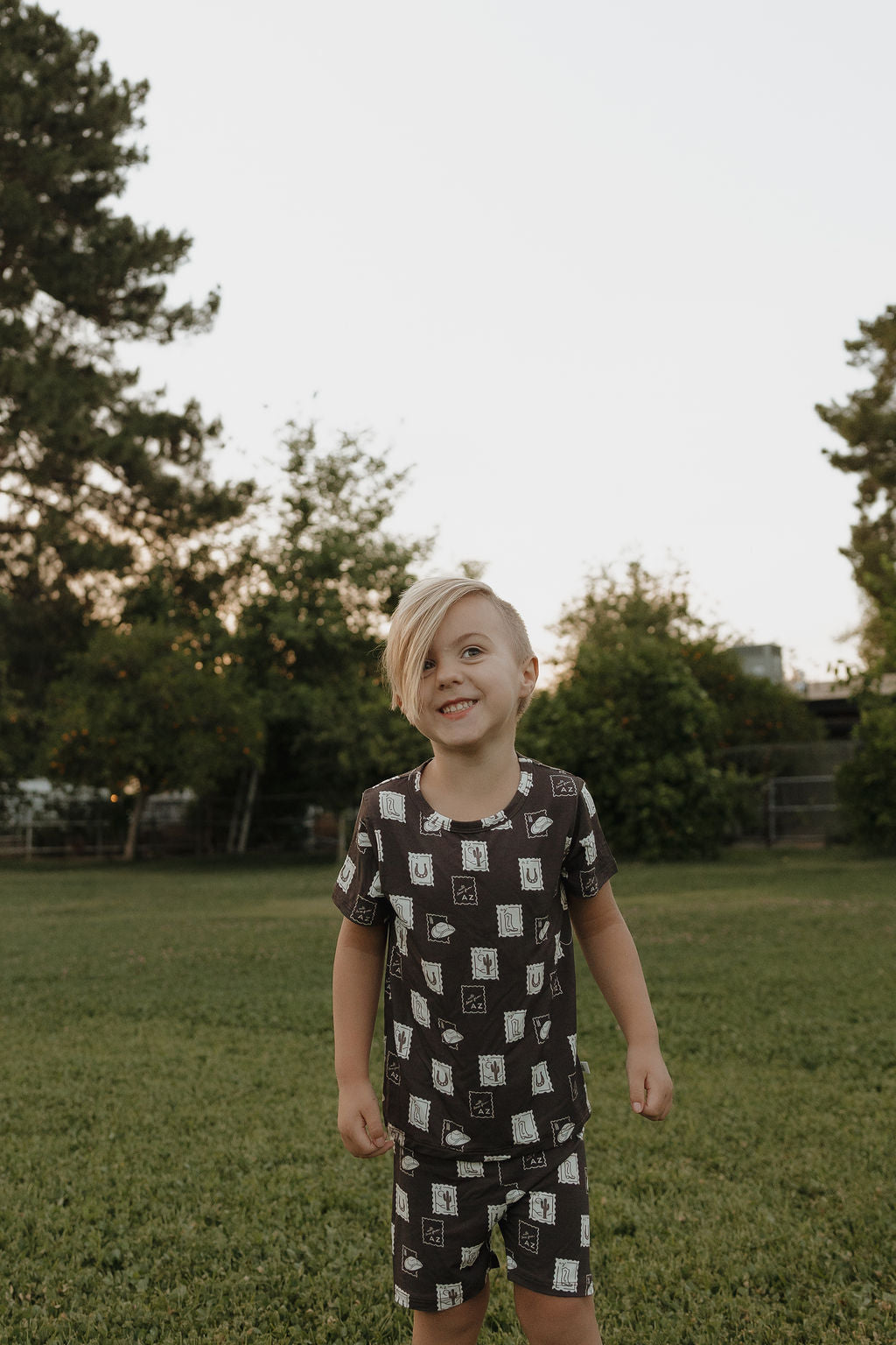 A young child with short blond hair smiles in a grassy field, wearing the forever french baby Bamboo Short Set | With Love from AZ—hypoallergenic kids clothing with a matching dark shirt and shorts. Trees and bushes can be seen in the background.