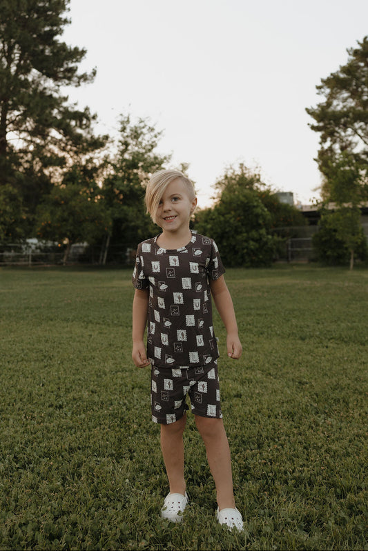 A young child with blonde hair smiles on a grassy field, wearing the forever french baby Bamboo Short Set | With Love from AZ—breathable matching shorts and T-shirt with light patterns and white slip-on shoes. Trees and greenery fill the background.
