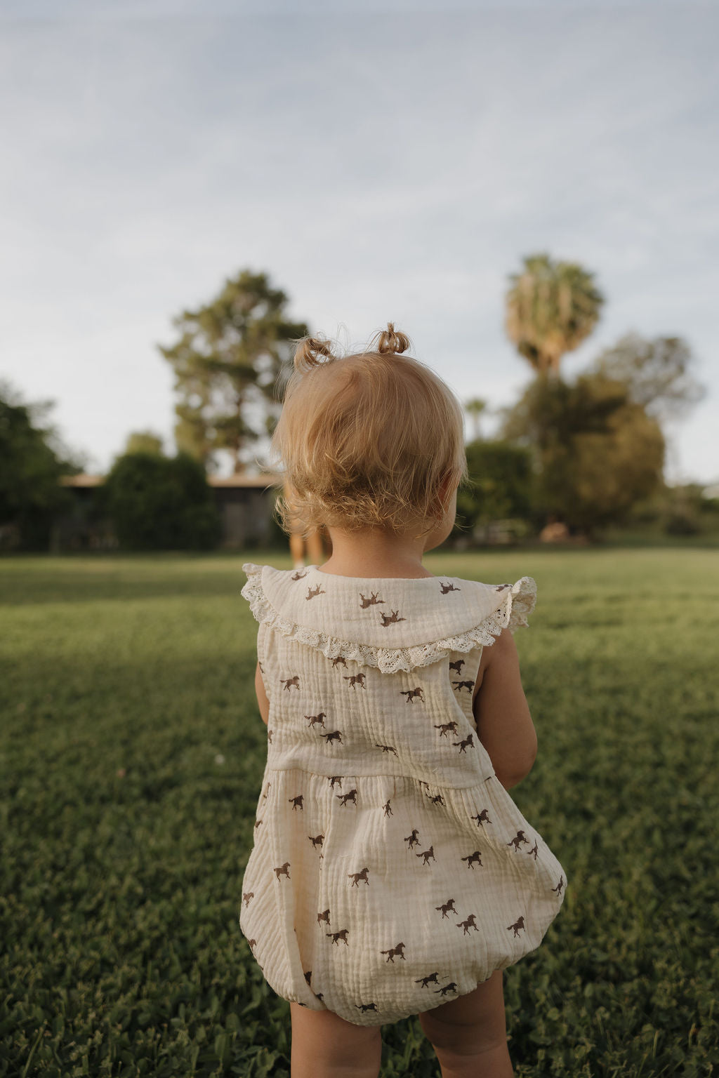 A young child with blonde pigtails stands on green grass, wearing the forever french baby Muslin Romper | Wild Horses. Trees and a house are in the background as she explores outdoors.