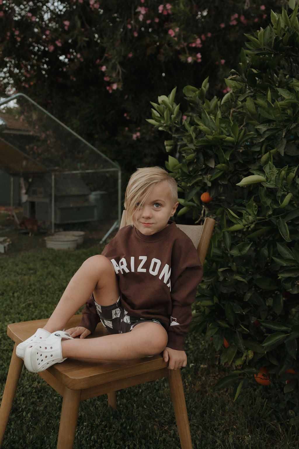 A young child with blonde hair, wearing the forever french baby Child Sweatshirt | ff Arizona, patterned shorts, and white shoes, sits on a wooden chair outside near an orange tree and garden structure.