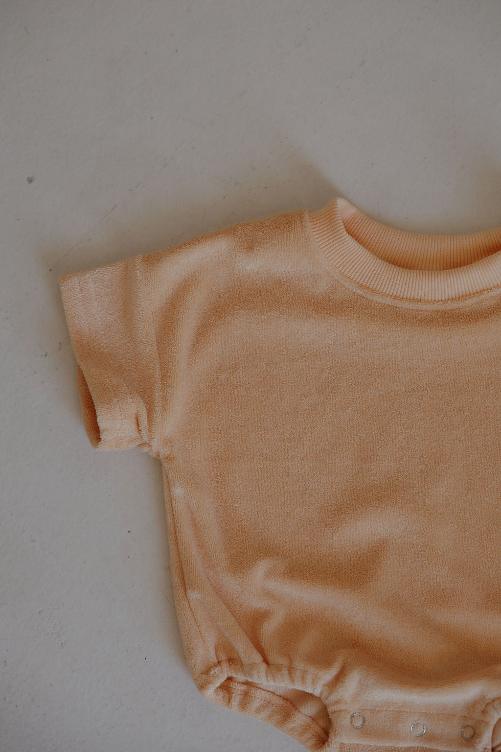 A close-up of the peach Sherbert Terry T-Shirt Romper by forever french baby, displayed flat to show short sleeves, the top half, and handy snap buttons at the bottom hem for easy diaper changes.