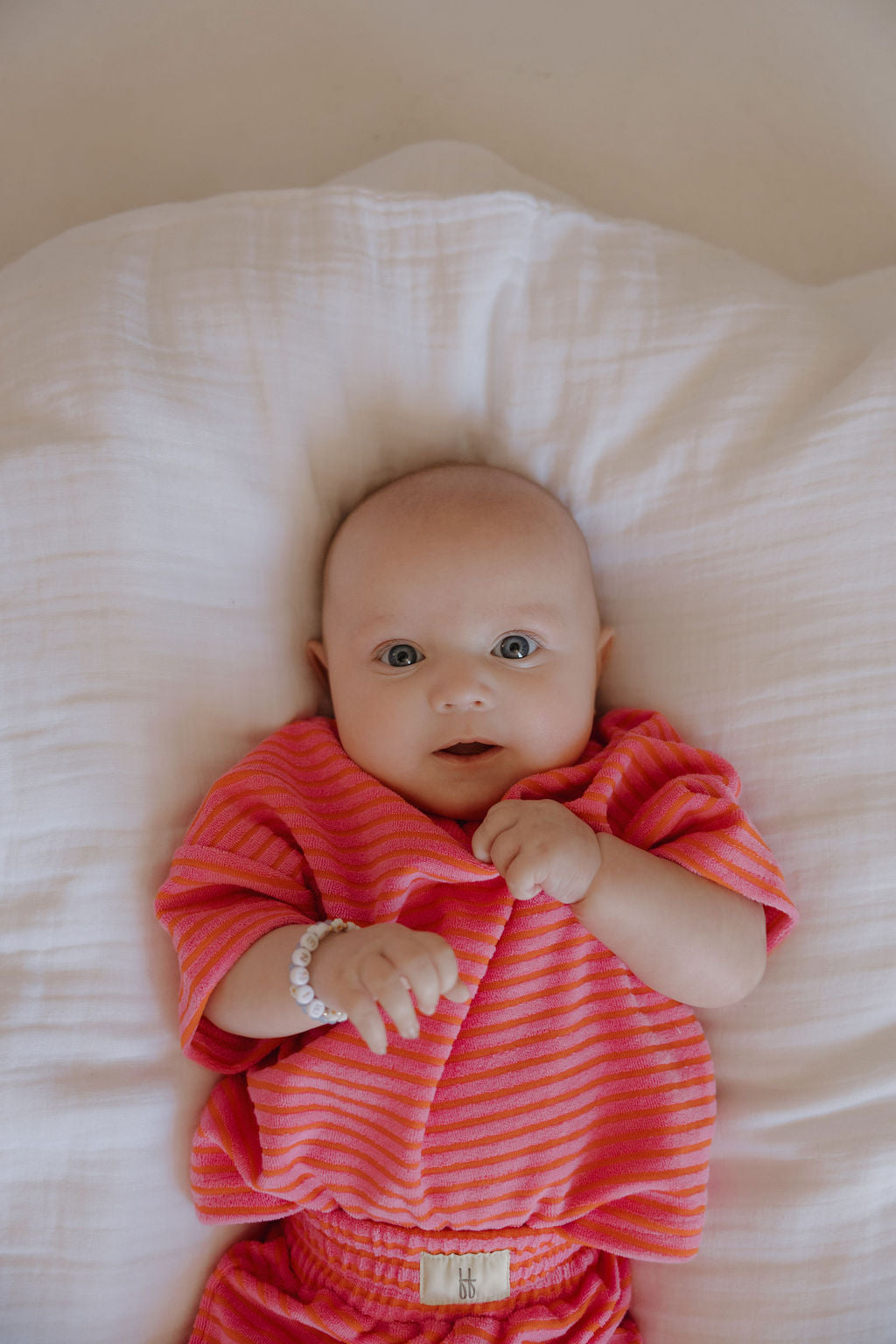 A baby with big eyes, dressed in the forever french baby Child Terry Short Set | Strawberry, lies on a white pillow with hands near their chest and wears a white beaded bracelet, capturing precious family moments.