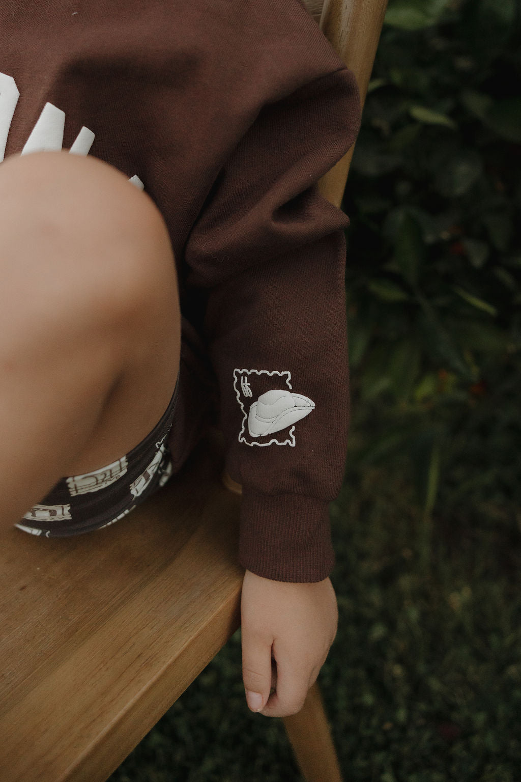 A young child sits on a wooden chair outdoors, wearing the forever french baby ff Arizona Child Sweatshirt with a white dinosaur graphic on the sleeve and patterned shorts. Green foliage can be seen in the background.