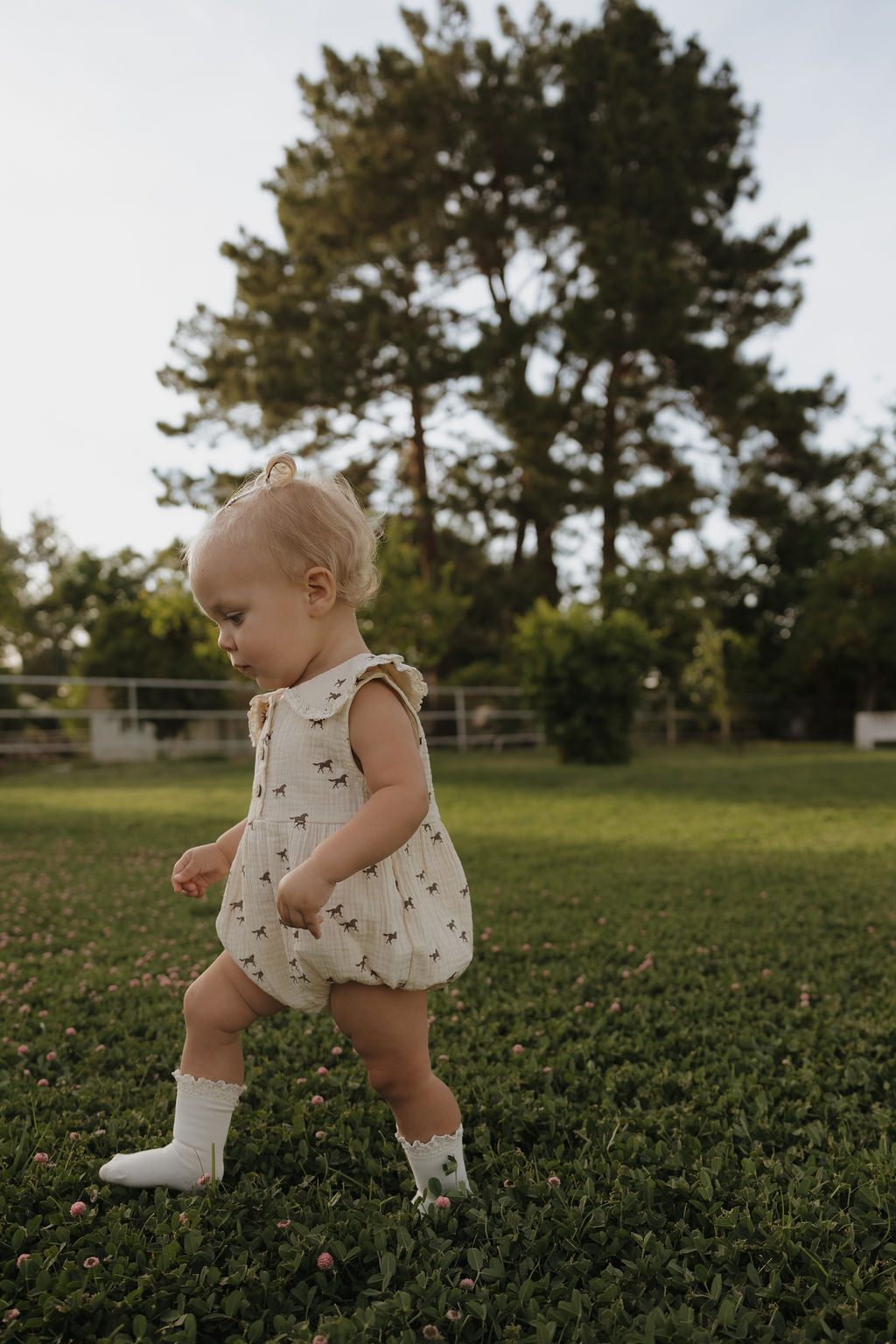 A young toddler with blonde hair, dressed in the forever french baby Muslin Romper | Wild Horses and white socks, walks on green grass outdoors with trees and a fence in the background.