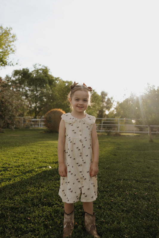 A young girl stands on green grass in a sunlit yard, wearing the forever french baby Child Muslin Dress | Wild Horses. Trees and a fence appear in the background, with sunlight shining from behind her.