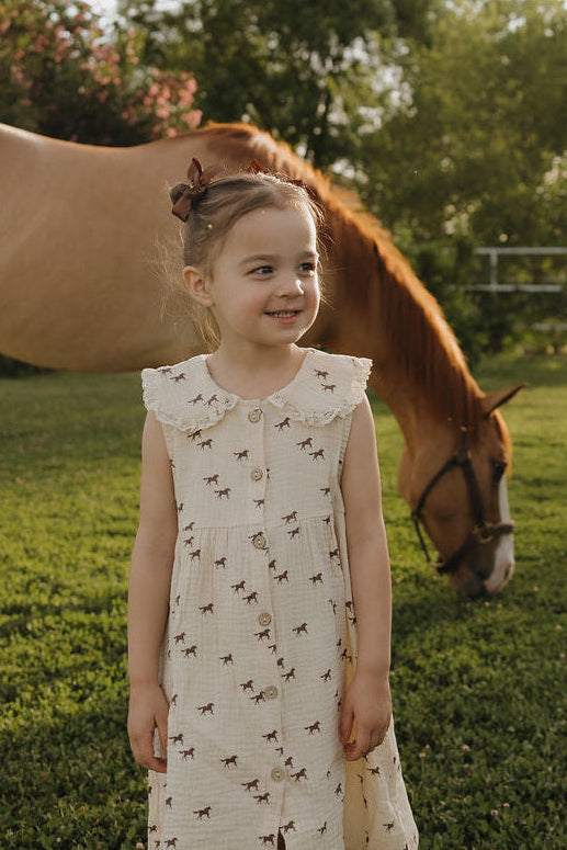 A young girl smiles in the forever french baby Child Muslin Dress | Wild Horses as she stands on grass, with a brown horse grazing behind her and sunlit trees in the background.