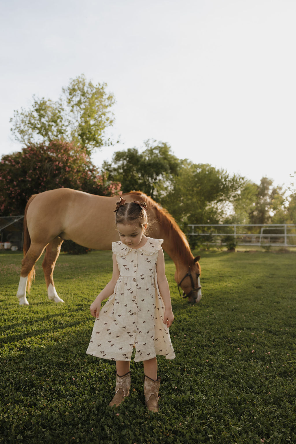 A girl wears the forever french baby Child Muslin Dress | Wild Horses, standing on green grass and looking down as a brown horse grazes behind her in a sunlit, tree-filled pasture.