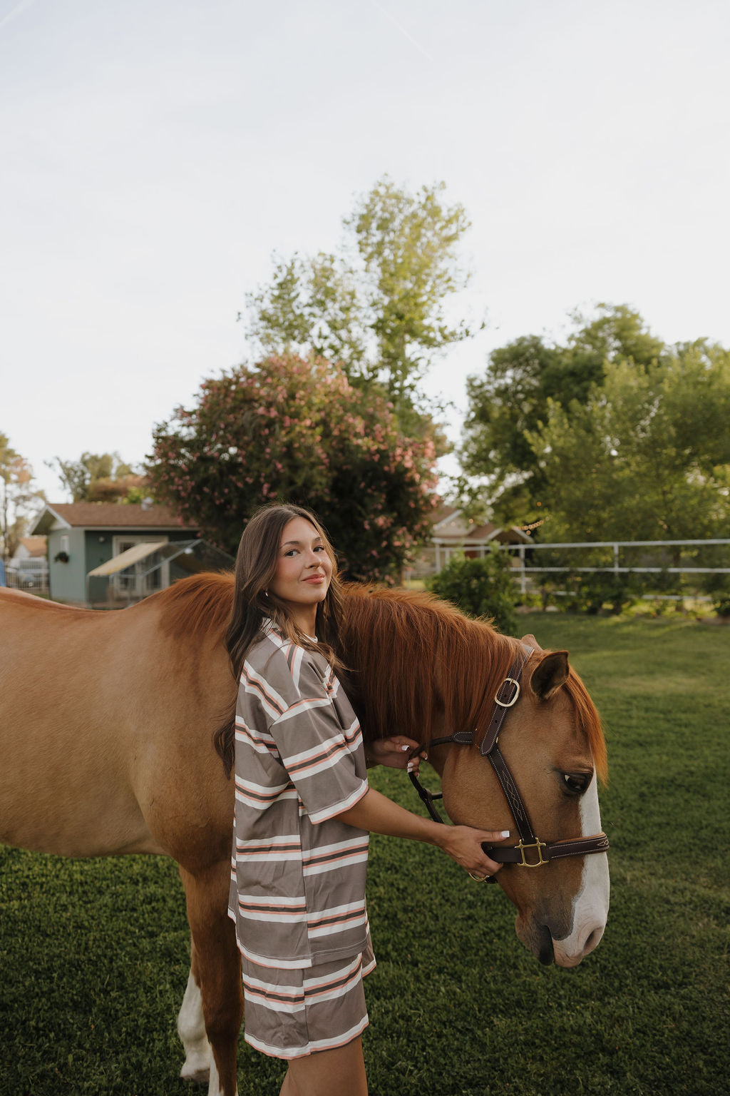 A woman in the forever french baby Women's Short Set | Sedona Stripe stands on green grass, gently holding a brown horse's bridle outdoors near trees and a house in soft evening light.