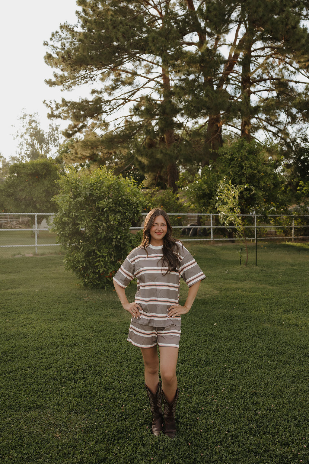 A woman with long hair stands on green grass in a yard, smiling with her hands on her hips. She wears the Women's Short Set | Sedona Stripe by forever french baby and dark boots, framed by trees, bushes, and a wire fence in the background.