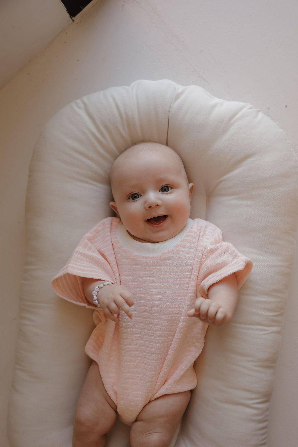 A smiling baby in the forever french baby Terry T-Shirt Romper | Cotton Candy lies on a soft, white cushion, looking up at the camera—perfect for capturing precious family moments.
