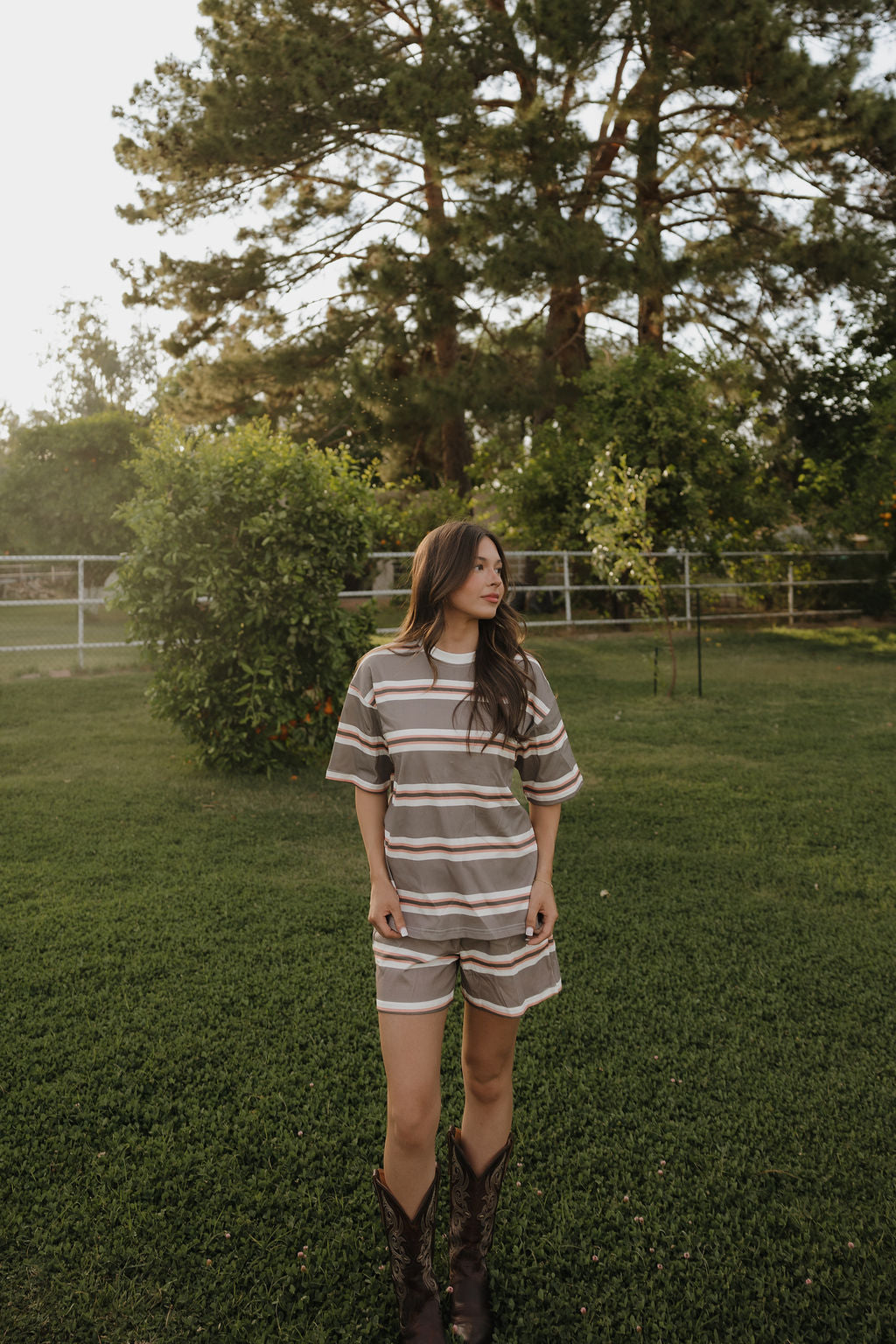 A woman in the forever french baby Women's Short Set | Sedona Stripe stands on grass in a park with tall trees and a fence behind her, styled with long hair and brown cowboy boots.