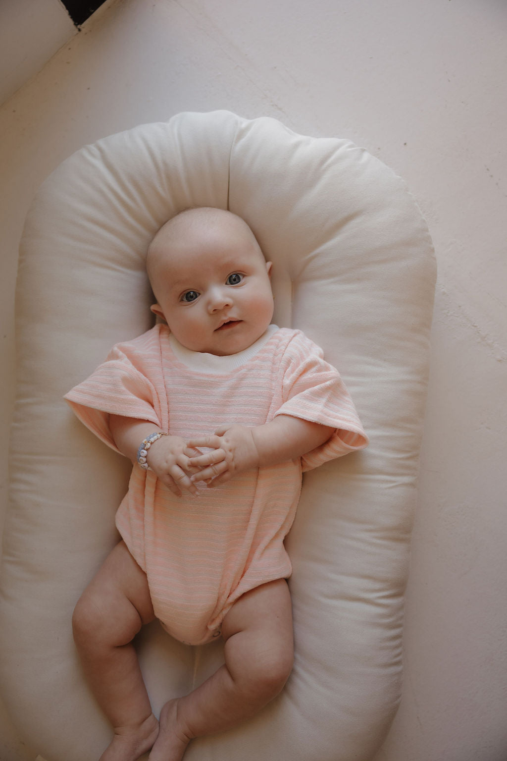 A baby in the forever french baby Terry T-Shirt Romper | Cotton Candy lies on a white cushioned pad, gazing up with hands together. The soft, minimal background highlights sweet family moments.