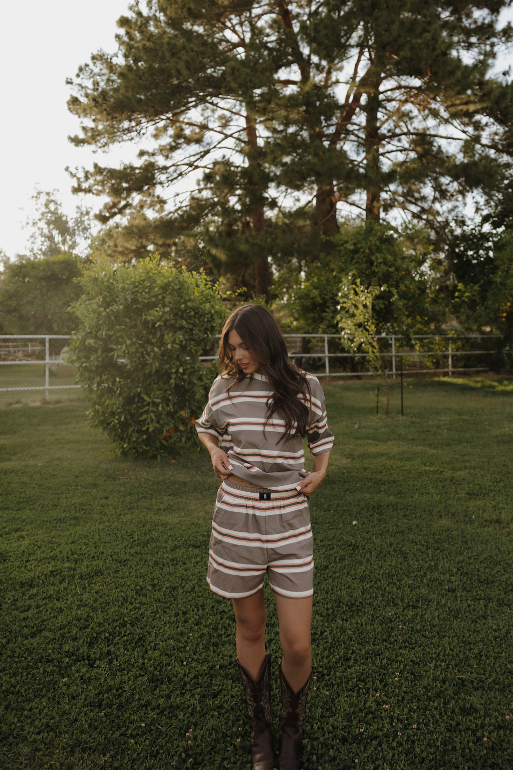 A woman with long brown hair stands on green grass, wearing the forever french baby Women's Short Set in Sedona Stripe and brown boots. She looks down with her hands in her pockets; trees and a fence are in the background.