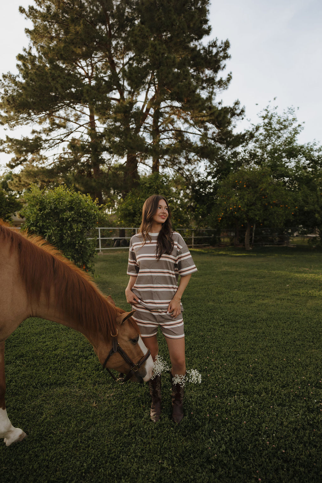 A woman wearing the forever french baby Women's Short Set in Sedona Stripe stands on green grass beside a brown horse, smiling and looking to the side with trees, a fence, and sunshine in the background.