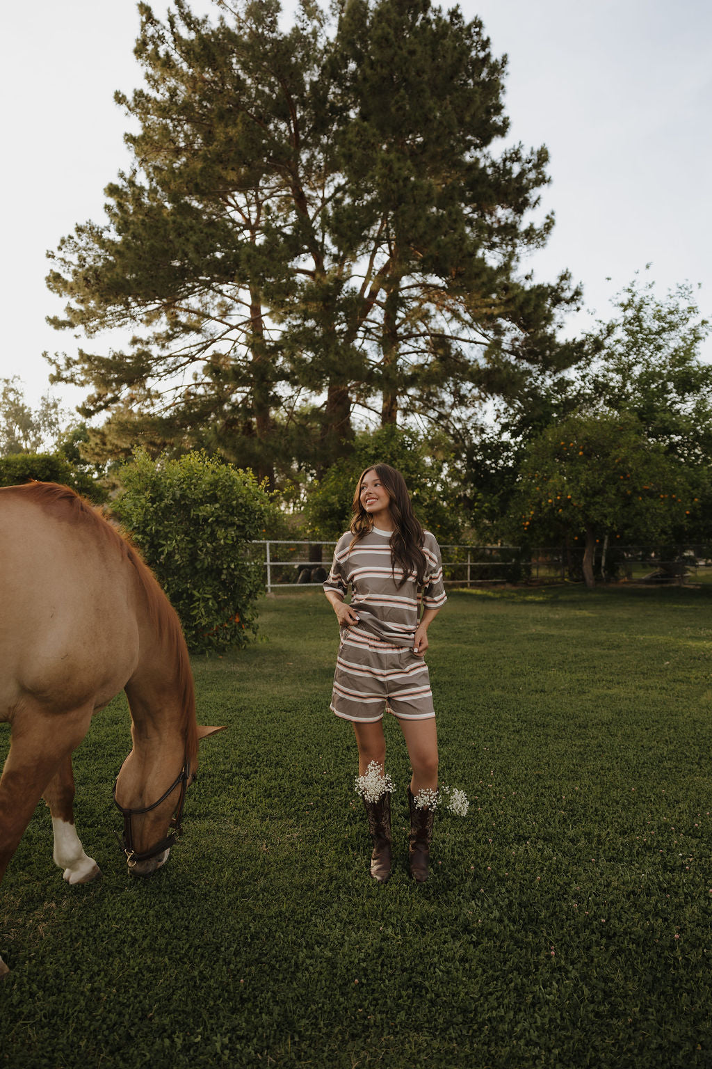 A woman in the Women's Short Set | Sedona Stripe by forever french baby and boots smiles on grass beside a brown horse grazing, with tall trees and a wooden fence in the background.