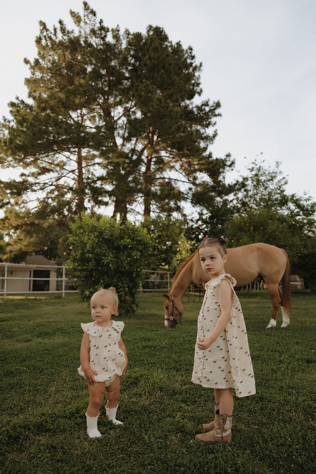 Two young girls in matching cream outfits stand on grass; one wears the forever french baby Child Muslin Dress | Wild Horses with cowboy boots, the other a romper and socks. A brown horse grazes nearby, framed by trees and a fence.
