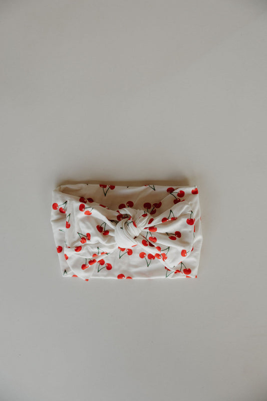 The Bamboo Head Wrap | Sweetheart Cherries from forever french baby features a white wrap with a central knot, decorated with red cherries and green stems—ideal for adding a charming touch to your little one’s outfit.