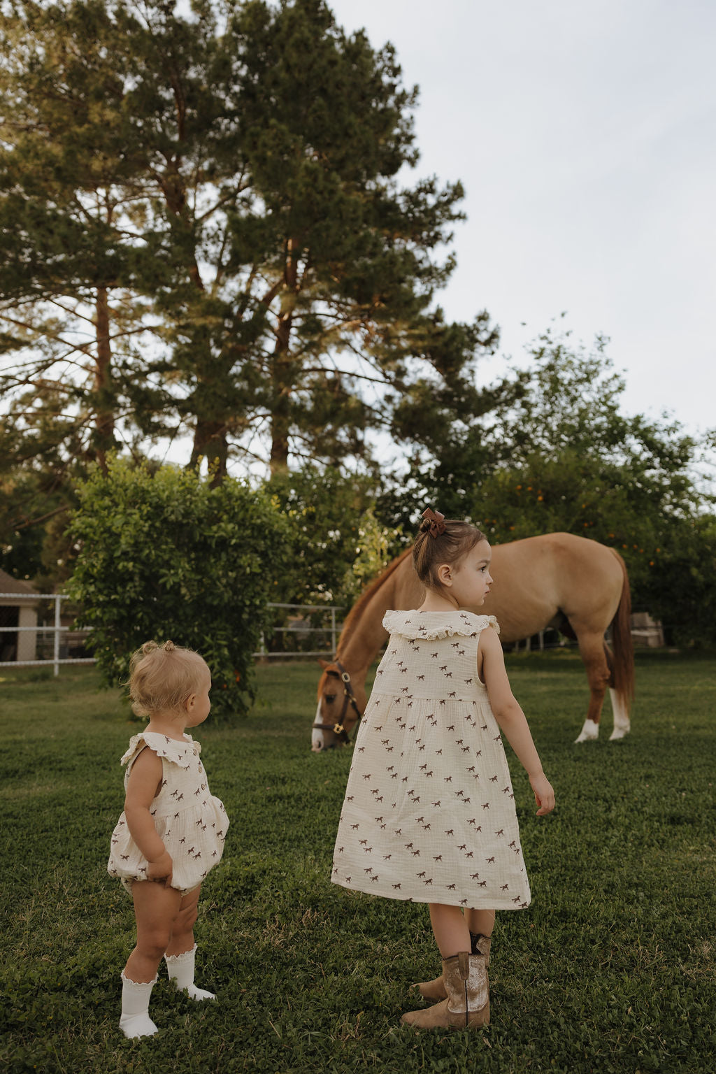 Two young children in matching outfits stand on green grass, watching a tan horse graze. One wears the forever french baby Child Muslin Dress | Wild Horses, blending perfectly with the peaceful outdoor scene.